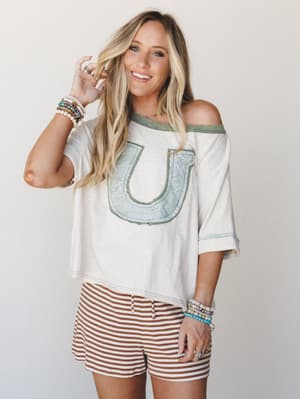 A relaxed, lived-in oversized tee with a horseshoe appliqué patch sits off one shoulder, paired with rust-striped shorts and layered boho bracelets — the kind of outfit that looks effortless because it genuinely is.