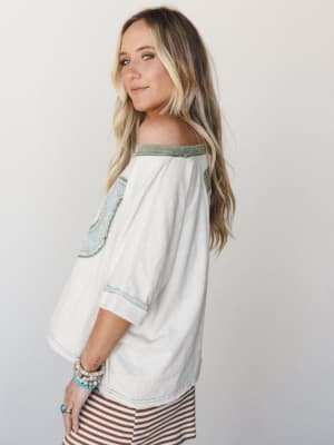 A relaxed off-shoulder top in cream with sage green contrast trim and scalloped lace-edged patch details, styled with a rust stripe skirt and stacked boho bracelets — the kind of top that feels like it has a good story.