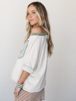 A relaxed off-shoulder top in cream with sage green contrast trim and scalloped lace-edged patch details, styled with a rust stripe skirt and stacked boho bracelets — the kind of top that feels like it has a good story.