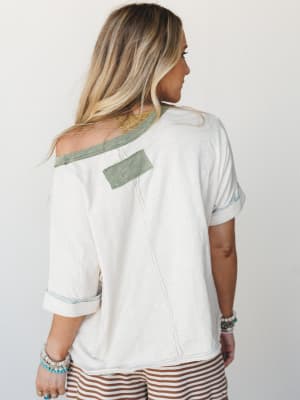 A back-view shot of a relaxed, oversized cream tee with sage green contrast trim and a fabric patch detail — styled with striped shorts and stacked boho bracelets for that effortlessly lived-in feel.