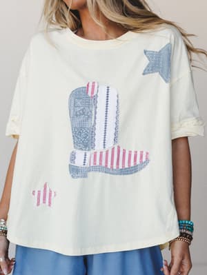A butter yellow oversized tee with a patchwork-style cowboy boot graphic and scattered stars in red stripe and bandana-print fabrics — worn with a blue skirt and stacked boho bracelets, it's got that effortless Americana-meets-Western energy.