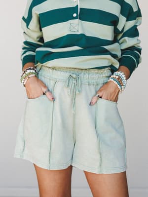 A relaxed mid-shot of acid-washed mint shorts paired with a bold green striped rugby top, hands tucked in pockets and stacked bracelets on both wrists — the kind of effortless outfit that looks like it came together without trying.