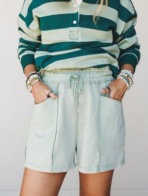 A relaxed mid-shot of acid-washed sage shorts paired with a bold green stripe polo, hands tucked in oversized patch pockets and stacked bead bracelets on both wrists — the kind of effortless outfit that feels intentional without trying.