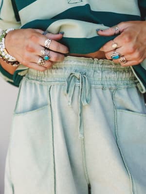 A close-up waist shot of washed sage green shorts with a drawstring waistband, styled with a teal stripe top and layered silver and turquoise jewelry — casual and collected with a distinctly boho hand.