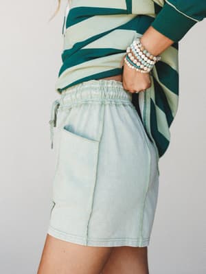 A close-up side shot of an easy, lived-in outfit — acid-washed mint shorts with a smocked waistband paired with a bold teal stripe top and a stack of beaded bracelets that feel collected, not coordinated.