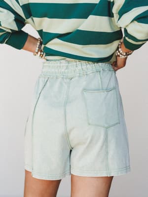 A back-view close-up of relaxed, washed-out sage shorts paired with a bold green stripe long-sleeve top — the kind of effortless, color-coordinated casual outfit that looks intentional without trying too hard.