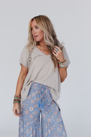 A woman with long blonde hair wears the Camden Lace V Neck Top in camel and blue floral wide-leg pants, accessorized with colorful bracelets and rings, standing against a plain light background.