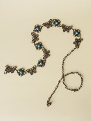 An antique brass chain belt laid loosely on a warm cream surface, its linked medallions alternating between filigree butterfly motifs and round turquoise cabochon flowers — the kind of piece that makes a simple dress feel like it has a story.