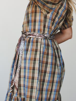 A close-up torso shot of a plaid midi dress cinched with a braided leather and paisley fabric belt — the kind of layered styling that looks effortless but clearly wasn't an accident.