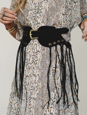 A close-up of a wide black suede fringe belt with brass buckle and grommet detail, styled over a printed cream dress with gold accents — the kind of accessory that does all the talking.