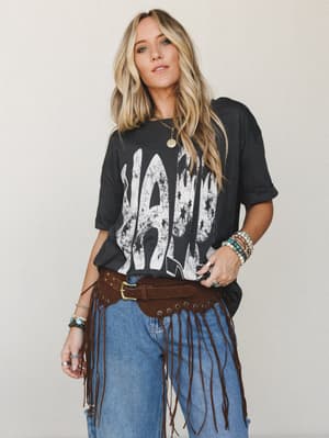 A lived-in graphic tee, worn-in denim, and a brown suede fringe belt styled together with stacked turquoise and gold jewelry — the kind of outfit that looks like it took five minutes but tells a whole story.