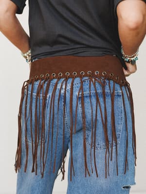 A back-view shot showing a wide brown suede fringe belt with brass grommet detailing worn over a black tee and light-wash denim jeans — the long leather fringe strands cascade mid-thigh and the stacked boho bracelets on both wrists pull the whole loo