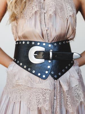 A wide black studded leather belt with an oversized silver western buckle cinches a blush lace dress at the waist — the contrast between soft and edgy is doing a lot of good work here.