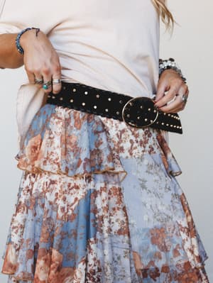 A close-up waist shot showing a black studded belt with a gold oval buckle layered over a floral tiered skirt and cream top — the kind of styling detail that pulls a whole look together.