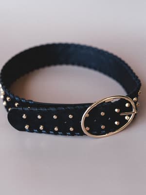 A black braided leather belt with scattered gold studs and an oval gold buckle, photographed close-up against a soft blush-grey background — the kind of finishing piece that makes a simple outfit feel intentional.