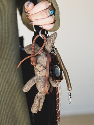 A close-up of a hand adorned with silver and turquoise rings and leopard-print nails, holding a woven mocha bunny bag charm alongside layered keychain accessories — the kind of detail shot that makes you want to raid someone's bag.