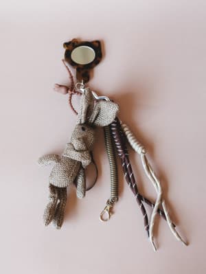 A whimsical bag charm collection laid flat on a blush background — centered on a crocheted bunny figure in mocha, paired with a tortoiseshell mirror charm, a coiled metal keyring, and braided rope straps in cream and burgundy.