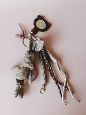 A crochet bunny bag charm in mocha laid flat on a dusty pink surface, styled alongside a coiled wristlet keychain, braided cords, and a tortoiseshell mini mirror — the kind of collected, character-filled accessories that make a bag feel personal.