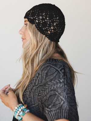 A profile shot of a blonde model wearing a black open-weave crochet cap paired with a charcoal cable-knit sweater top and a stack of beaded bracelets — earthy and effortless with just enough edge.