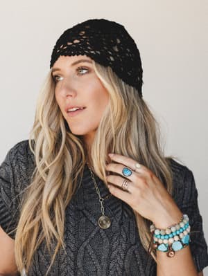 A close portrait of a model wearing a black crochet beanie over loose waves, layered with a cable-knit sweater, stacked turquoise beaded bracelets, stone rings, and a gold coin pendant necklace — the kind of effortlessly collected look that feels int