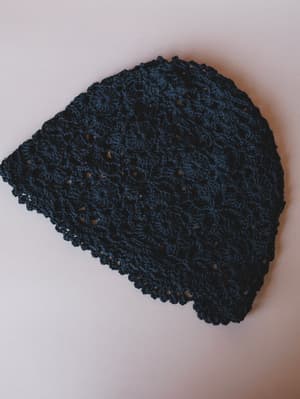 A deep navy-black crochet beanie laid flat on a neutral background, showing off its open floral stitch pattern and scalloped edge — the kind of hat that makes a simple outfit feel intentional.