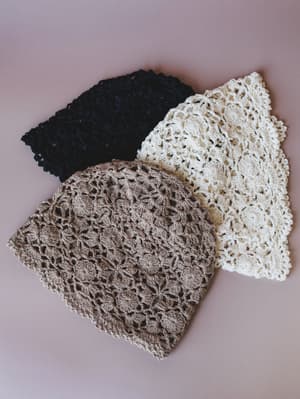 Three crochet beanies laid flat in a loose fan arrangement — taupe, cream, and black — each showing the same open floral stitch pattern with scalloped edges, shot on a muted dusty rose surface.