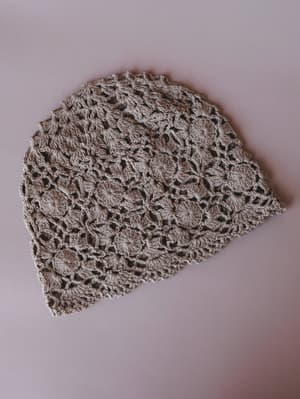 A mocha-toned crochet beanie laid flat against a soft blush-pink surface, its open floral stitch pattern giving it a handcrafted, vintage-market feel.