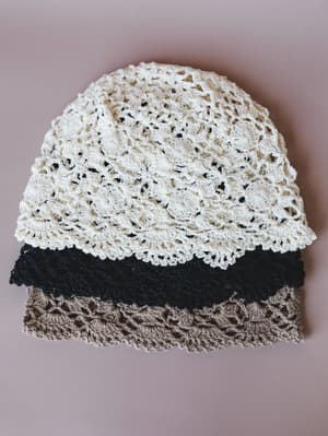 Three stacked crochet beanies — cream, navy, and mocha — photographed flat against a dusty mauve background, showing off the open-stitch shell pattern and scalloped edges in a clean, simple product shot.
