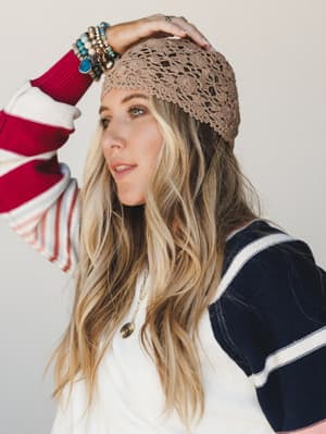 A close-up portrait of a model wearing a mocha crochet beanie with a delicate open-stitch pattern, styled casually with a striped color-block sweater and layered jewelry — effortlessly boho, like she just grabbed the hat on the way out the door.