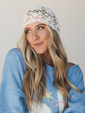 A close-up portrait of a model wearing a delicate cream crochet cap paired with a soft blue graphic sweater — the kind of effortless boho pairing that looks like it just happened.