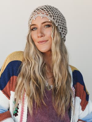 A close-up portrait of a model wearing a delicate beige crochet net headband with pearl embellishments, layered over a colorful striped knit cardigan and a dusty mauve waffle-knit top — the whole look feels effortlessly boho with a soft, vintage-girl