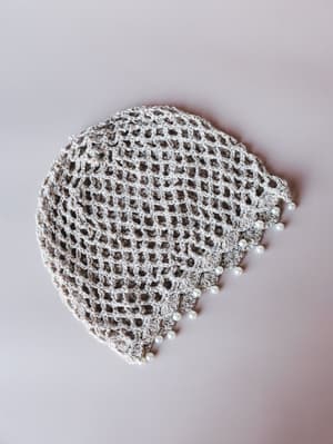 A crochet net hair snood in soft beige, laid flat against a blush-grey background — delicate open weave finished with pearl bead trim along the edge, equal parts vintage and wearable.