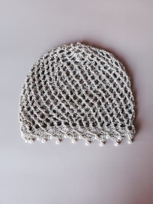 A flat lay of an open-weave crochet beanie in soft beige, finished along the edge with dangling pearl beads — the kind of piece that feels equally at home at a festival or a bridal shower.