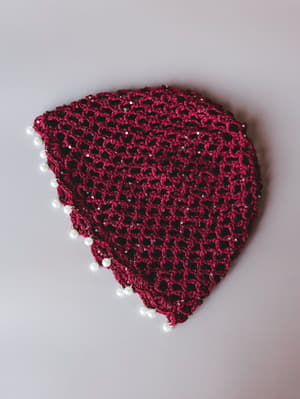 A triangular crochet headband in deep wine red, laid flat against a soft gray backdrop — the open net stitch catches scattered sequins throughout, while pearl beads line the edges for a detail that feels unexpectedly special.