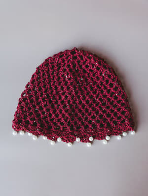 A wine-colored crochet beanie laid flat, its open-weave mesh catching tiny scattered sequins throughout and finished along the brim with dangling pearl beads — the kind of piece that makes a simple outfit feel like a decision.