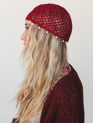 A side-profile shot of a model wearing a deep wine crochet net beanie adorned with dangling pearl embellishments along the brim, paired with a burgundy boucle sweater and a floral layer peeking underneath — the kind of hat that makes a whole outfit f