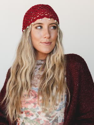 A close-up portrait of a blonde model wearing a deep wine crochet net headband with pearl bead fringe, layered over a floral top and burgundy fuzzy cardigan — boho romantic with just enough sparkle to feel special.