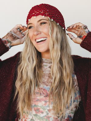 A laughing model adjusts a cranberry red crochet headband trimmed with pearl drops, layered over a floral mesh top and a deep burgundy fuzzy cardigan — the whole look has that effortless 'threw it together and it worked' energy.