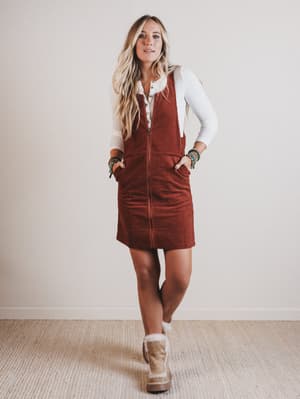 Woman wearing a brown corduroy pinafore dress with a white shirt underneath, standing against a plain background.