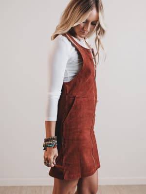 Woman wearing a brown corduroy pinafore dress over a white shirt against a plain background