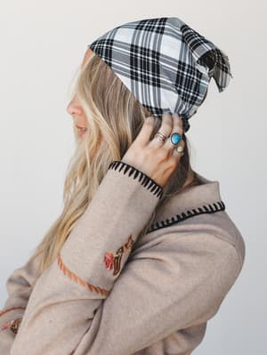 A side-profile close-up of a woman tying a black and white plaid bandana into her hair, her stacked turquoise and silver rings catching the light against a soft oat-toned embroidered sweater — the whole thing feels like a Saturday morning with great