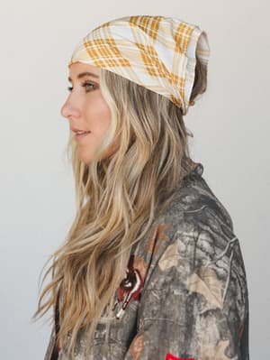A side-profile shot of a model wearing a slouchy mustard plaid beanie-style bandana, paired with a woodland camo jacket with embroidered detail — casual and a little unexpected in the best way.
