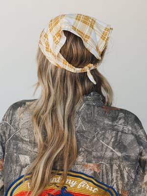 Shot from behind, a model wears a mustard plaid bandana tied at the back of her head over a camo jacket with embroidered back patch detailing — casual, a little vintage, very much her own thing.