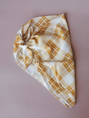 A mustard and cream plaid bandana tied into a knot, laid flat against a muted dusty rose background — the kind of easy accessory that pulls a whole look together without trying.