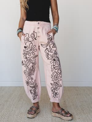 Blush pink jogger-style pants covered in bold black floral embroidery, styled with a black crop top, beaded bracelets, and espadrille platform sandals — the kind of statement bottom that does all the talking.