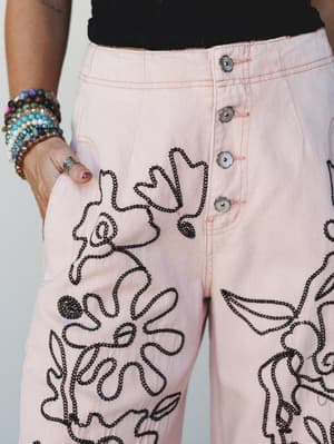 A close-up of high-waisted blush denim pants with bold black chain-stitch floral and botanical embroidery covering the legs, styled with a black crop top and stacked beaded bracelets — the kind of pants that do all the talking.