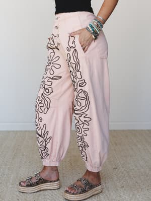 Blush pink wide-leg jogger pants with bold black abstract embroidery cascade down both legs, styled with a black tank, stacked turquoise bracelets, and leopard print platform sandals — effortlessly expressive and ready for wherever the day takes her.