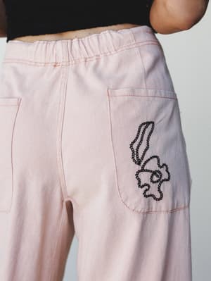 A close-up detail shot of blush pink wide-leg pants with contrast pink topstitching, patch pockets, an elasticized waist, and a playful hand-stitched floral embroidery motif on the hip — the kind of detail you notice and immediately want to point out