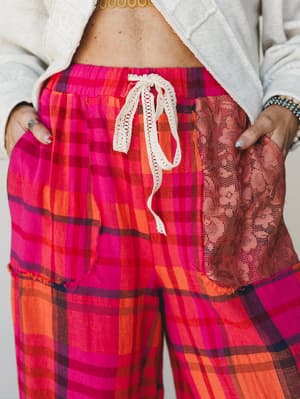 A close-up detail shot of bold pink and orange plaid wide-leg pants with a crochet drawstring waist and a floral lace patch pocket — the kind of pants that make the whole outfit.