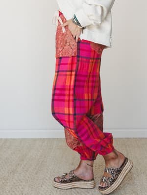 A side-profile shot of bold pink plaid harem-style pants with lace patchwork panels, styled with a cream sweatshirt, stacked bracelets, and leopard print platform sandals — expressive and comfy in equal measure.
